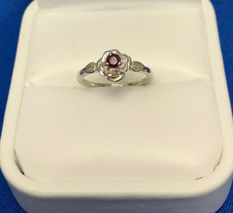 May include: A silver ring with a red gemstone set in a rose design. The ring is in a white ring box.