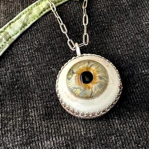 May include: A silver chain necklace with a pendant shaped like a human eye. The eye is white with a brown iris and a black pupil. The pendant is surrounded by a silver beaded border.