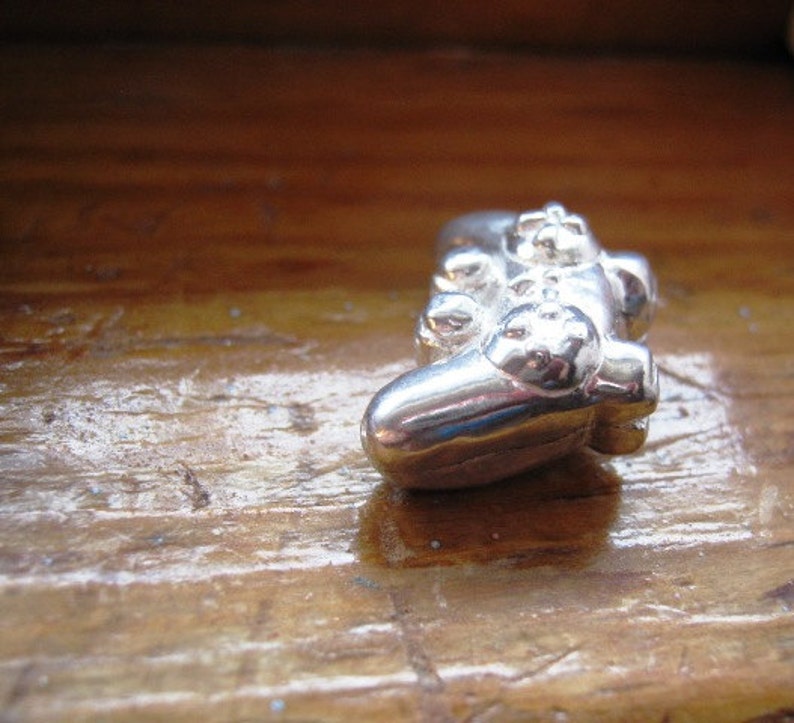 May include: A silver-colored, miniature replica of a video game controller. The controller is lying on a wooden surface.