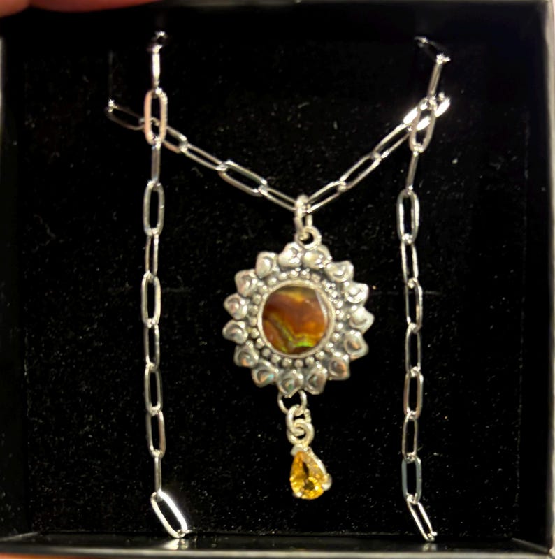 K&ouml;nnte beinhalten: Eine silberne Halskette mit einem Anh&auml;nger, der einen runden, schillernden Stein zeigt, der von einem silbernen Blumendesign umgeben ist. Ein kleinerer, tropfenf&ouml;rmiger Stein h&auml;ngt an der Blume.