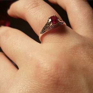 Pode incluir: Um anel de prata com uma pedra preciosa vermelha redonda e brilhante. O anel tem um design de filigrana ornamentado na banda. O anel está num dedo.