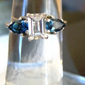 Puede incluir: Un anillo de plata con una piedra central grande, clara, de corte esmeralda, flanqueada por dos piedras más pequeñas, azules y de forma de pera.