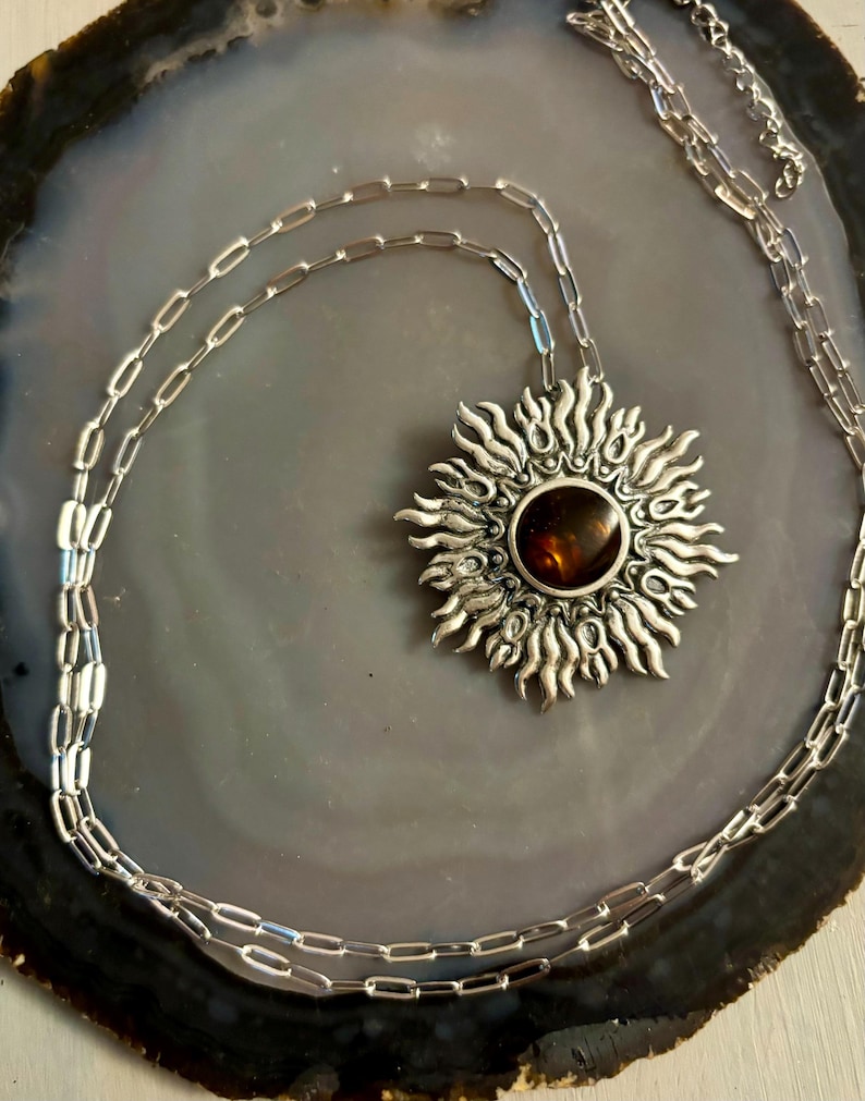 K&ouml;nnte beinhalten: Eine silberne Halskette mit einem Sonnenanh&auml;nger, der mit einem braunen Edelstein verziert ist. Die Kette ist in einem zarten, verlinkten Stil.