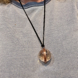 May include: A clear crystal ball pendant with a copper band around the middle, hanging from a black cord necklace.