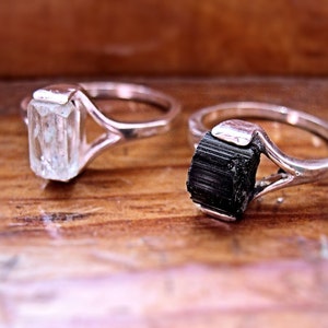 May include: Two silver rings with gemstone settings. The ring on the left has a clear, faceted gemstone. The ring on the right has a black, striated gemstone.