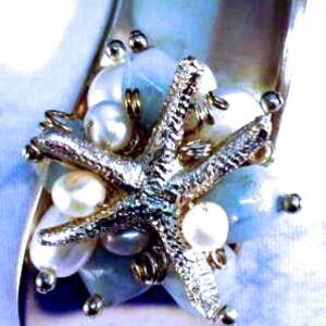 May include: A silver starfish charm with white pearls and blue stones, set on a white shell background. The charm is a beautiful and unique piece of jewelry.