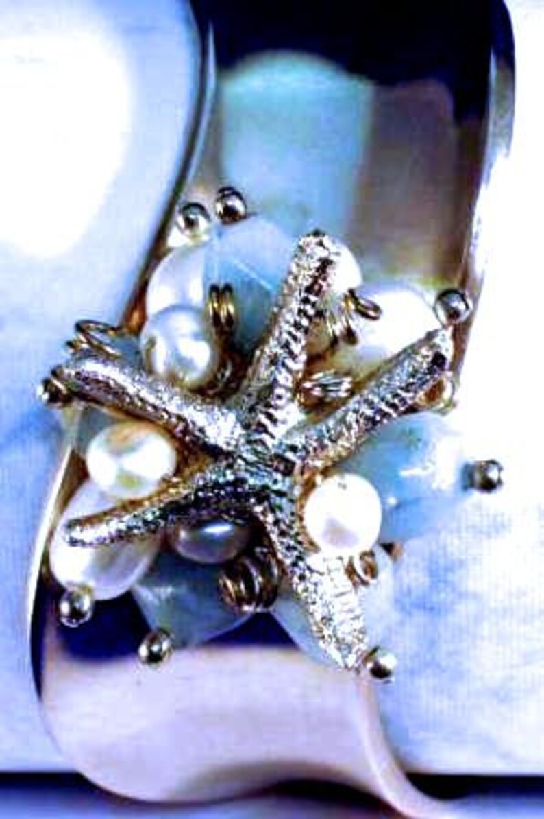 May include: A silver starfish charm with white pearls and blue stones, set on a white shell background. The charm is a beautiful and unique piece of jewelry.