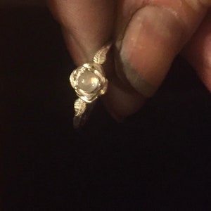 May include: A silver ring with a clear gemstone set in a rose-shaped design. The ring has delicate leaf accents on either side of the stone.