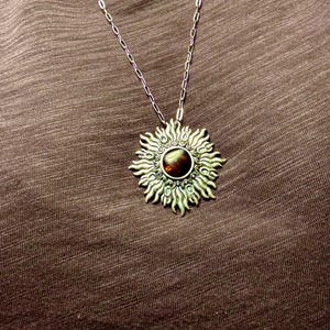 K&ouml;nnte beinhalten: Eine silberne Halskette mit einem gro&szlig;en Sonnenanh&auml;nger, der einen braunen Edelstein in der Mitte tr&auml;gt. Der Anh&auml;nger hat ein detailliertes Design mit filigranen Strahlen, die vom Zentrum ausgehen.
