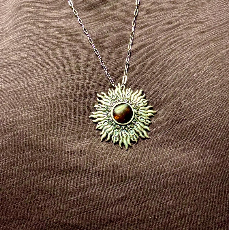 K&ouml;nnte beinhalten: Eine silberne Halskette mit einem gro&szlig;en Sonnenanh&auml;nger, der einen braunen Edelstein in der Mitte tr&auml;gt. Der Anh&auml;nger hat ein detailliertes Design mit filigranen Strahlen, die vom Zentrum ausgehen.