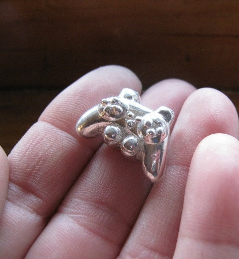 May include: A silver-colored pendant shaped like a video game controller. The pendant has a detailed design with buttons and a joystick.