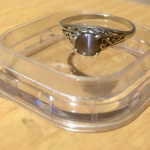 May include: A silver ring with a gray cat's eye gemstone set in a filigree design. The ring is displayed in a clear plastic ring box.