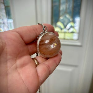 Può includere: Una collana con pendente in argento con una grande gemma rotonda e traslucida con inclusioni marrone-rossastre. La gemma è incastonata in una montatura d'argento decorativa e pende da una delicata catena d'argento. Il pendente è tenuto in una mano.