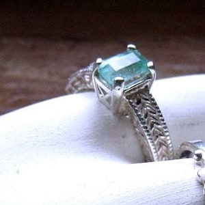 May include: A silver ring with a light green emerald-cut gemstone set in a delicate filigree design.