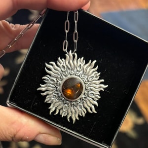 K&ouml;nnte beinhalten: Eine silberne Halskette mit einem Sonnenanh&auml;nger, der einen braunen Edelstein in der Mitte tr&auml;gt. Der Anh&auml;nger ist mit komplizierten Designs verziert und hat eine strukturierte Oberfl&auml;che.