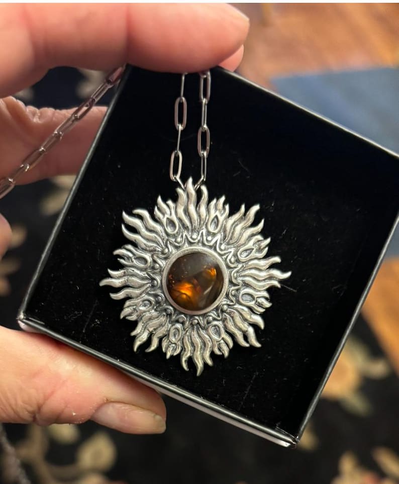 K&ouml;nnte beinhalten: Eine silberne Halskette mit einem Sonnenanh&auml;nger, der einen braunen Edelstein in der Mitte tr&auml;gt. Der Anh&auml;nger ist mit komplizierten Designs verziert und hat eine strukturierte Oberfl&auml;che.
