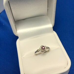 May include: A silver ring with a red gemstone set in a rose design. The ring is in a white ring box.