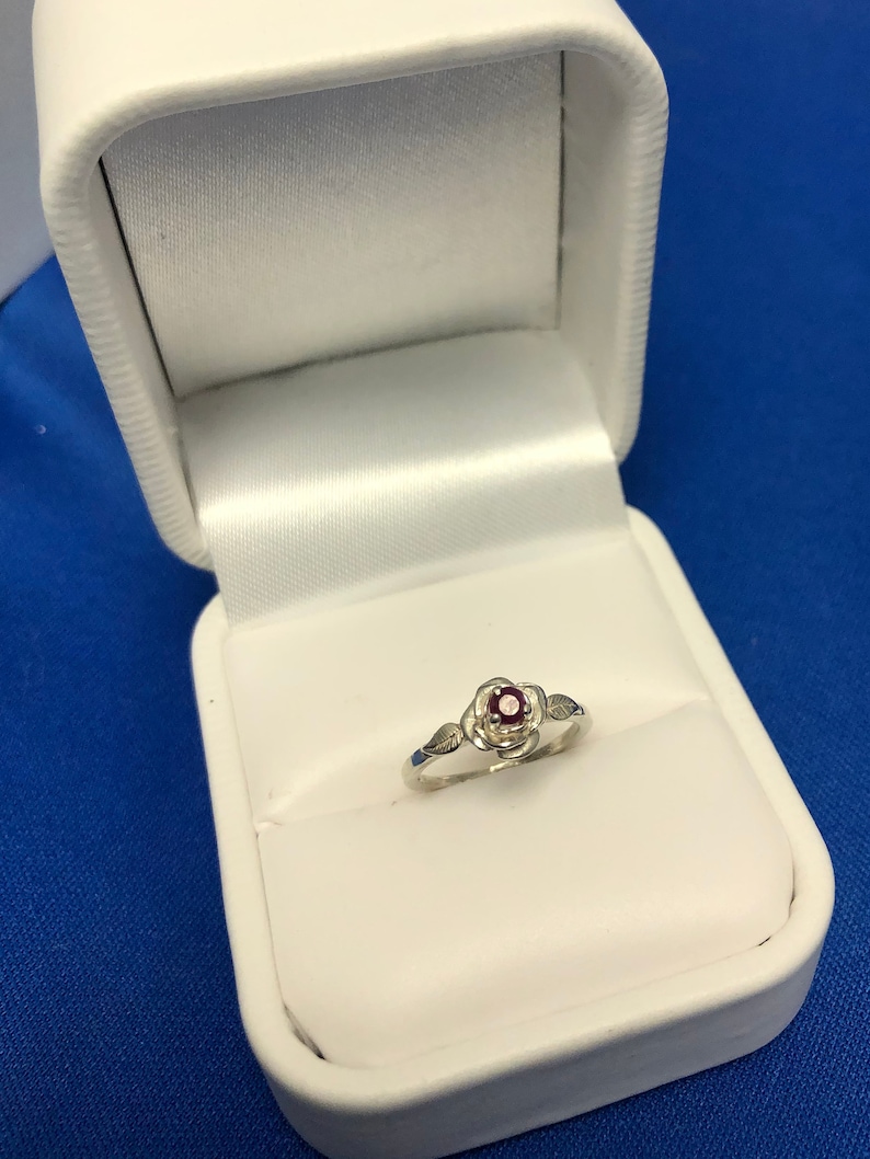 May include: A silver ring with a red gemstone set in a rose design. The ring is in a white ring box.