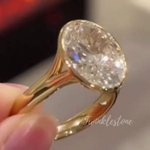 May include: A gold ring with a large, oval-cut diamond. The ring has a delicate, split-shank design. The diamond sparkles with many facets. The text "Twinklestone" is visible on the ring's band.