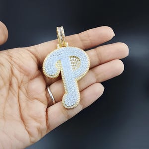 May include: A gold-toned pendant in the shape of the letter "P", encrusted with small, sparkling white stones. The pendant has a bail at the top for a chain. The letter is outlined in gold and held in a hand.