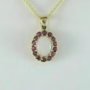 May include: A gold-colored pendant necklace featuring an oval, white opal stone surrounded by a halo of small, round, dark red gemstones. The pendant hangs from a delicate gold chain.