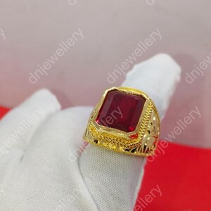May include: A gold-colored ring with a large, rectangular, red gemstone. The ring has intricate detailing around the gemstone and on the band. The ring is displayed on a white surface.