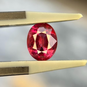 May include: A vibrant, oval-cut gemstone with a deep red hue and multifaceted surface, held securely between the tips of a pair of light-colored precision tweezers. The stone displays internal light reflections and a clear, polished finish.