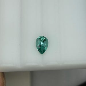 May include: A teal-coloured, pear-shaped gemstone with multiple facets. The gem is set against a white background, highlighting its clarity and brilliance. The gemstone's cut allows light to reflect, creating a sparkling effect.