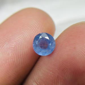 May include: A round, faceted, light blue gemstone. The gem is held between two fingers, showcasing its clarity and color. The stone has a polished surface and a brilliant cut, reflecting light.