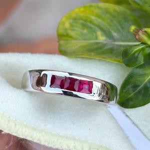 May include: A silver ring featuring three rectangular, vibrant red gemstones. The ring has a polished, reflective surface, and the gemstones are set in a row. The background includes green leaves and a soft, light-colored fabric.