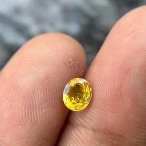 May include: A vibrant, oval-cut yellow gemstone. The gem is held between fingers, showcasing its brilliant facets and sunny color. The stone's clarity and polished surface reflect light, highlighting its brilliance.