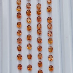 May include: A collection of oval-cut, orange-yellow gemstones arranged in neat rows. The gems are displayed on a white, ridged surface, showcasing their polished facets and warm color. These gemstones are ideal for jewelry making.