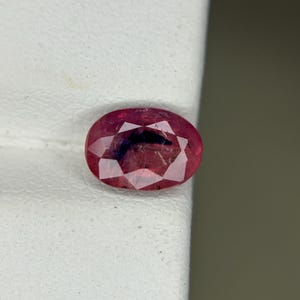 May include: A faceted, oval-shaped ruby gemstone. The gem is a deep red color with internal inclusions, and it is set against a white background. The cut of the stone creates multiple facets that reflect light.