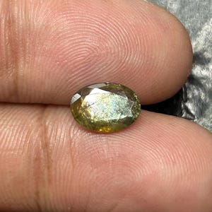 May include: An oval-cut gemstone with a mix of yellow and green hues. The gem has a faceted surface that reflects light, creating a shimmering effect. The stone is held between two fingers, showcasing its size and shape.