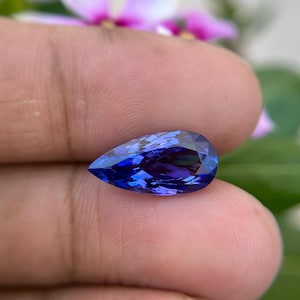 Puede incluir: Una gema facetada en forma de pera, de color azul intenso. Las facetas de la gema reflejan la luz, creando un efecto brillante. La piedra se sostiene entre los dedos, con un fondo floral borroso.