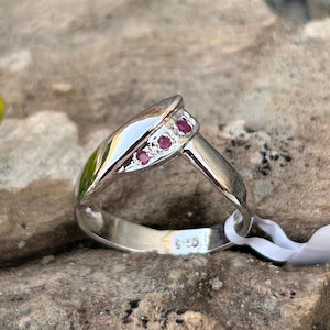 Könnte beinhalten: Ein silberner Ring mit geschwungenem Design, besetzt mit drei kleinen roten Edelsteinen. Der Ring hat eine polierte Oberfläche und ist vor einem natürlichen, strukturierten Hintergrund platziert. Der Ring ist ein Schmuckstück.