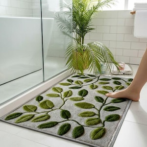 May include: A grey rectangular bath mat featuring a green leaf design. The leaves, in various shades of green, are arranged in a vine pattern. The mat is positioned in front of a shower or bathtub, measuring approximately 60cm x 90cm.