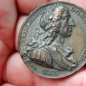 Puede incluir: Una moneda de bronce con el perfil de un hombre con cabello rizado y una corona de laurel. La moneda tiene inscripciones, incluyendo la palabra "REX". La moneda está sostenida en una mano.