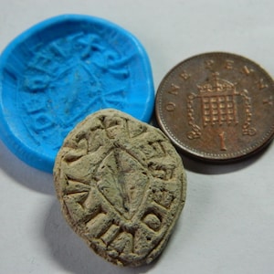 Puede incluir: Un sello de piedra desgastada con un diseño en forma de diamante y texto grabado, junto a un molde azul y una moneda de un penique de bronce. Los objetos están sobre un fondo claro.