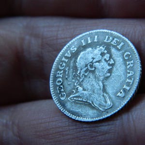 Puede incluir: Una moneda de plata con la inscripción "GEORGIVS III DEI GRATIA". La moneda muestra el perfil de una persona. La moneda se sostiene en una mano.