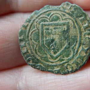 Puede incluir: Un artefacto circular de bronce desgastado con un diseño central de escudo. El escudo está rodeado por un borde ornamental, similar a flores. El color general es verde apagado con áreas marrones, sugiriendo antigüedad y exposición.