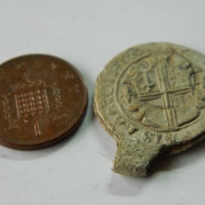 Puede incluir: Una moneda de bronce de un penique junto a un sello gris circular y desgastado. El sello presenta un diseño de cruz dentro de un borde circular. La moneda es un penique.