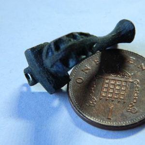 Puede incluir: Un pequeño objeto metálico oscuro con forma cilíndrica y base cuadrada, junto a una moneda de un penique. La moneda es de color bronce y muestra el número 1 y las palabras "ONE PENNY". El objeto parece ser una antigüedad o una escultura en miniatura.