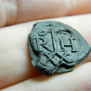 Puede incluir: Un objeto circular oscuro y desgastado con las letras "RH" y otras marcas. El objeto parece estar hecho de un material oscuro, posiblemente metálico. Las letras están en relieve y el diseño general es intrincado.