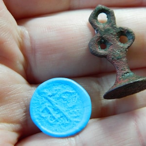 May include: An antique bronze seal with a decorative handle and a round base, next to a bright blue wax impression. The seal has a detailed design and shows signs of age and wear. The wax impression has a detailed design.