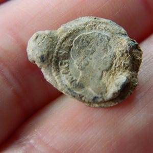 Puede incluir: Un pequeño objeto metálico circular y desgastado con un perfil en relieve de una persona y texto parcial. El objeto es de color gris claro y parece ser un sello o ficha antigua.