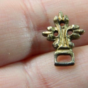 Puede incluir: Un pequeño dije de metal dorado y ornamentado. El dije tiene una parte superior decorativa con tres aberturas circulares y una base rectangular con una sola abertura. El dije está en la mano de una persona.