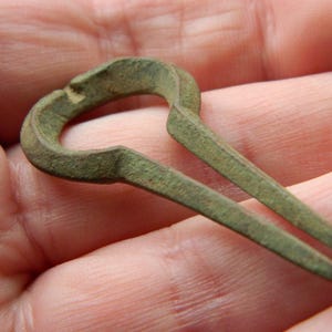 Puede incluir: Un arpa de boca de metal de tono bronce desgastado, con un marco circular y dos puntas paralelas que se extienden desde la base. La superficie del instrumento muestra una pátina envejecida y texturizada con signos visibles de oxidación y desgaste.