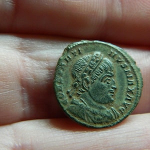 Op de afbeelding: Een kleine, ronde, antieke bronzen munt. De munt toont een profiel van een persoon met een kroon, omringd door tekst. De munt heeft een verweerd uiterlijk, met een groene patina.