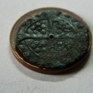 Puede incluir: Una moneda pequeña y oscura, posiblemente antigua, con un diseño detallado, apoyada sobre una moneda de color cobre. La superficie de la moneda es texturizada y desgastada, con un agujero central. El diseño parece ser una cruz o un patrón simétrico similar.